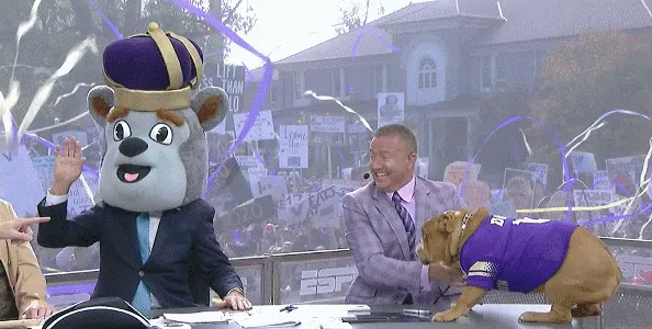 Lee Corso wearing the Duke Dog mascot's head waving whilst scaring a bulldog sitting on a desk.